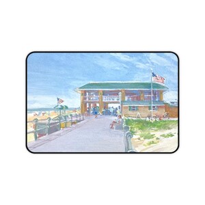 May include: A watercolor painting of a coastal boardwalk leading to a multi-level wooden building with a green roof. An American flag flies from a pole near the structure. The scene includes a blue sky, sandy areas, and figures on the walkway.
