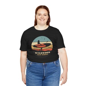 Puede incluir: Camiseta negra con un dise&ntilde;o gr&aacute;fico circular que representa a un surfista con una tabla de surf y el texto "Wildwood New Jersey". El dise&ntilde;o utiliza una combinaci&oacute;n de colores de atardecer con tonos naranja, amarillo y azul. La camiseta la lleva una persona con vaqueros azules.