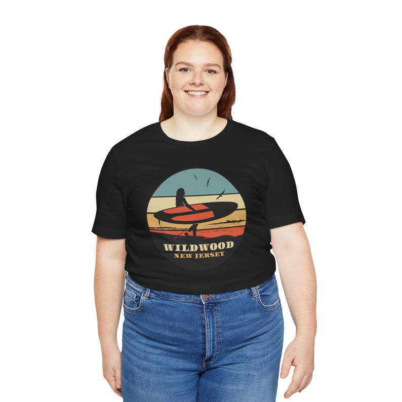 Puede incluir: Camiseta negra con un dise&ntilde;o gr&aacute;fico circular que representa a un surfista con una tabla de surf y el texto "Wildwood New Jersey". El dise&ntilde;o utiliza una combinaci&oacute;n de colores de atardecer con tonos naranja, amarillo y azul. La camiseta la lleva una persona con vaqueros azules.
