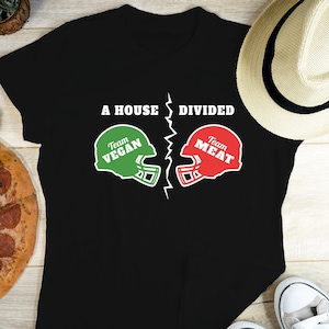 May include: Black t-shirt with the text "A HOUSE DIVIDED" above two football helmets. One helmet is green with "Team VEGAN", the other is red with "Team MEAT". A straw hat and a white shoe are partially visible.