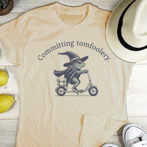 May include: Beige t-shirt with a whimsical illustration of a frog riding a scooter, wearing a witch hat and cape. The text "Committing tomfoolery" arches above the frog. A straw hat and sneakers are visible.