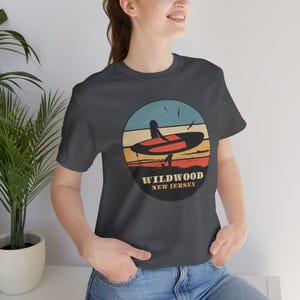 Puede incluir: Camiseta gris oscuro con un dise&ntilde;o gr&aacute;fico circular que muestra una tabla de surf y el texto "Wildwood New Jersey". El dise&ntilde;o incluye una persona con una tabla de surf en una playa con un fondo de puesta de sol. La camiseta es de material suave.