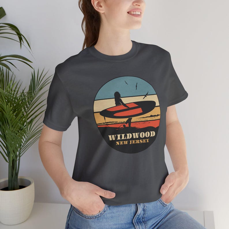 Puede incluir: Camiseta gris oscuro con un dise&ntilde;o gr&aacute;fico circular que muestra una tabla de surf y el texto "Wildwood New Jersey". El dise&ntilde;o incluye una persona con una tabla de surf en una playa con un fondo de puesta de sol. La camiseta es de material suave.