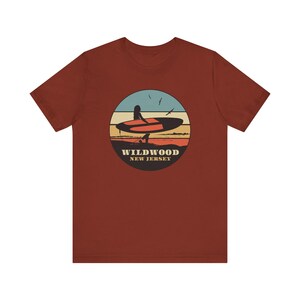 Puede incluir: Camiseta de color &oacute;xido con un dise&ntilde;o gr&aacute;fico circular de una persona en una tabla de surf al atardecer. El dise&ntilde;o incluye un cielo azul, rayas naranjas y amarillas y el texto "WILDWOOD NEW JERSEY".