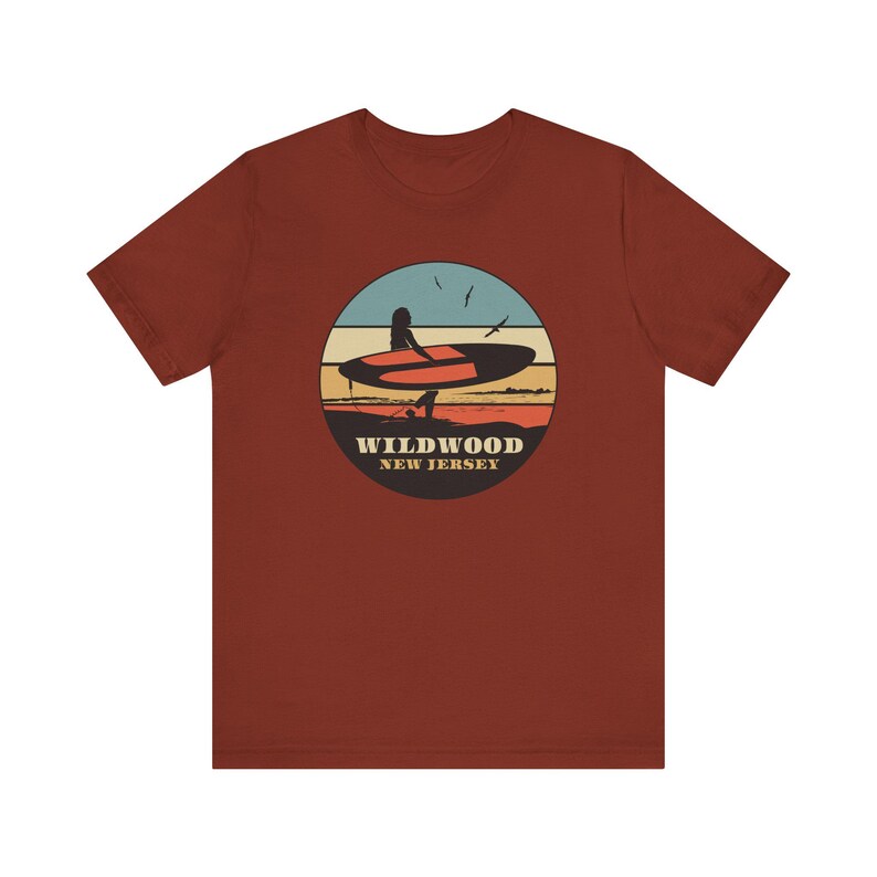 Puede incluir: Camiseta de color &oacute;xido con un dise&ntilde;o gr&aacute;fico circular de una persona en una tabla de surf al atardecer. El dise&ntilde;o incluye un cielo azul, rayas naranjas y amarillas y el texto "WILDWOOD NEW JERSEY".