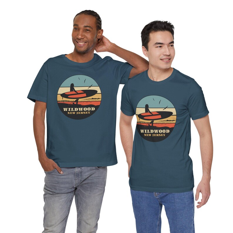 Puede incluir: Dos camisetas de color verde azulado con un gr&aacute;fico circular que presenta una tabla de surf, una persona y el texto "Wildwood New Jersey". El gr&aacute;fico tiene un dise&ntilde;o retro de puesta de sol con tonos naranja, amarillo y azul. Las camisetas son de manga corta.