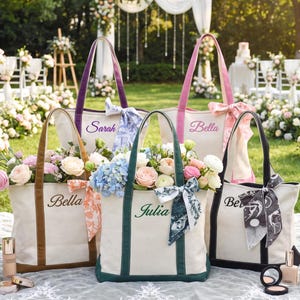 May include: Five off-white canvas tote bags with colored handles and bases in purple, pink, brown, dark green, and black. Each bag features embroidered names including Sarah, Bella, and Julia, and is decorated with a patterned silk scarf tied to one handle.