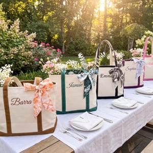 Könnte beinhalten: Fünf Tragetaschen aus Segeltuch auf einem Tisch, jede mit unterschiedlich farbigen Rändern und gesticktem Namen: Barra in Braun, Jamie in Grün, Emma in Schwarz, Sophia in Lila und Melin in Pink, verziert mit gemusterten Seidenbändern.