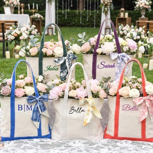 May include: Five off-white canvas tote bags with colored handles and bases in blue, green, purple, and red. Each bag is personalized with names or initials in script or block lettering and features a decorative patterned ribbon tied to one handle.