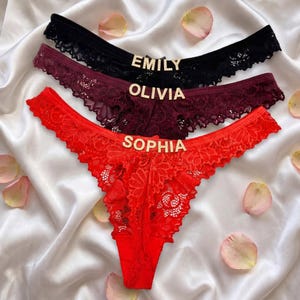 May include: Three pairs of lace thong underwear in black, burgundy, and red. Each pair features a gold-toned metal nameplate centered on the waistband, displaying the names EMILY, OLIVIA, and SOPHIA respectively, with floral lace detailing.