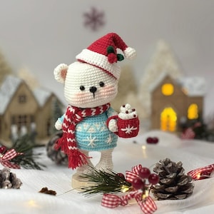 May include: A crocheted polar bear figurine wearing a red and white Santa hat and scarf, and a blue sweater with snowflakes. The bear holds a red mug with whipped cream. Christmas-themed decor includes miniature houses, pinecones, and ribbons.