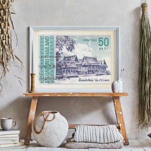 Op de afbeelding: Een vintage-print van een Thais huis met een groene en witte rand. De print is ingelijst in een licht houten lijst en hangt aan een lichtgrijze muur. De print is omringd door verschillende woonaccessoires, waaronder een geweven mand, een keramische vaas en een stapel boeken.