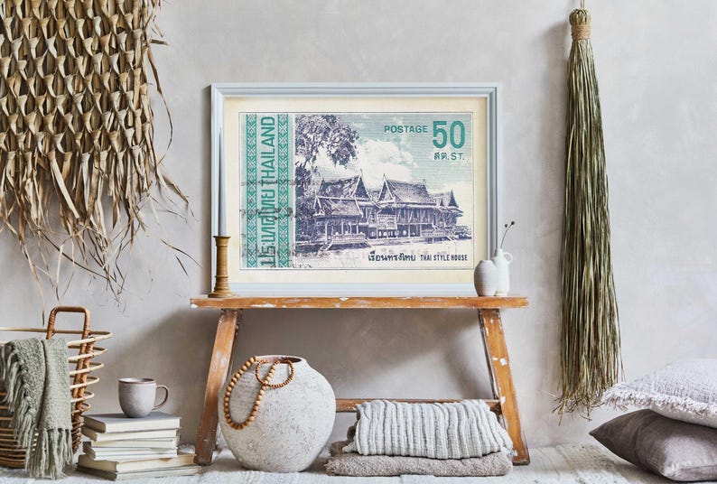 Op de afbeelding: Een vintage-print van een Thais huis met een groene en witte rand. De print is ingelijst in een licht houten lijst en hangt aan een lichtgrijze muur. De print is omringd door verschillende woonaccessoires, waaronder een geweven mand, een keramische vaas en een stapel boeken.