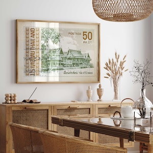 Op de afbeelding: Een ingelijste print van een vintage Thaise postzegel met een traditioneel Thais huis met groen gebladerte en de tekst "15:ทศไทย THAILAND POSTAGE 50 สต.ST. เรือนทรงไทย THAI STYLE HOUSE". De print hangt aan een witte muur boven een houten kast met geweven rotan deuren.