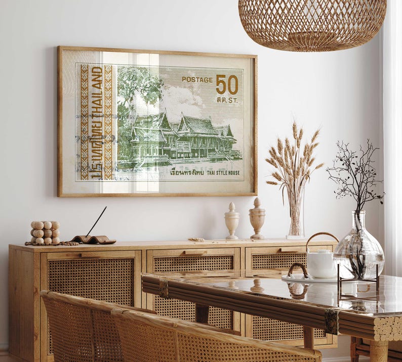 Op de afbeelding: Een ingelijste print van een vintage Thaise postzegel met een traditioneel Thais huis met groen gebladerte en de tekst "15:ทศไทย THAILAND POSTAGE 50 สต.ST. เรือนทรงไทย THAI STYLE HOUSE". De print hangt aan een witte muur boven een houten kast met geweven rotan deuren.