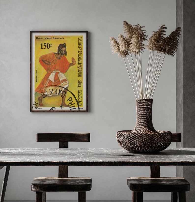 Pu&ograve; includere: Un poster vintage incorniciato con sfondo giallo che presenta una donna con un vestito rosso e bianco che balla. Il poster &egrave; intitolato "Kono - danse Bamanan" e "Republique du Mali" con il testo "150F" nell'angolo in alto a sinistra. Il poster &egrave; appeso a una parete bianca sopra un tavolo di legno con due sedie.