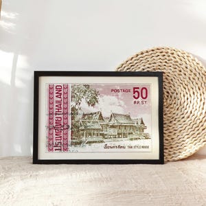 Op de afbeelding: Een ingelijste vintage Thaise postzegel met een rode rand en de tekst "ประเทศไทย THAILAND" en "POSTAGE 50 สด.ST." De postzegel toont een gedetailleerde illustratie van een traditioneel Thais huis.