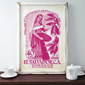 May include: A framed vintage El Salvador postage stamp print in shades of pink and white. The stamp features a woman holding a basket, surrounded by botanical elements and text. The print is displayed on a dark wooden surface.