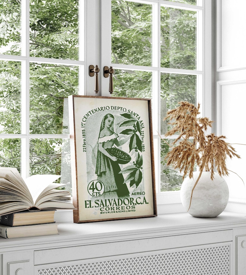 May include: A framed vintage El Salvador stamp, featuring a woman holding a basket, with text in Spanish. The stamp is green and brown, set against a window with a white frame and a view of green foliage.