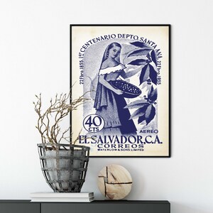 May include: A framed vintage poster with a blue and white design. The poster features a woman holding a basket, with text that reads "EL SALVADOR, C.A. CORREOS" and "CENTENARIO DEPTO. SANTA ANA". A decorative plant and wooden sculpture are on a dark cabinet.