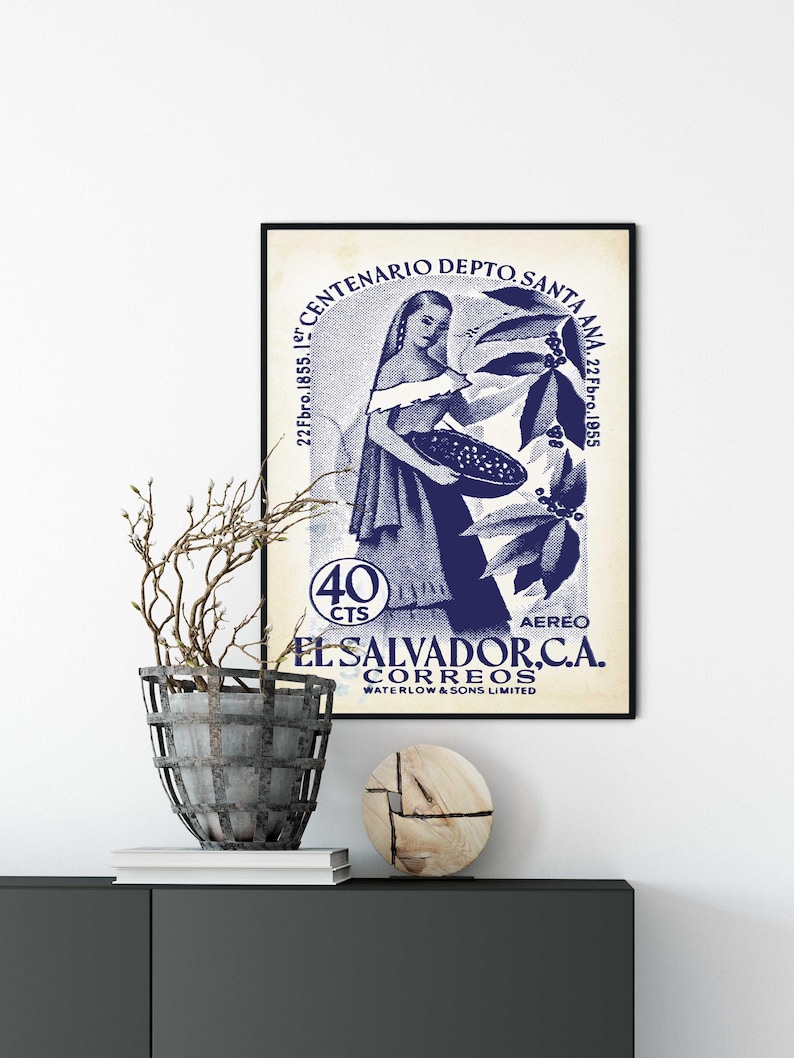 May include: A framed vintage poster with a blue and white design. The poster features a woman holding a basket, with text that reads "EL SALVADOR, C.A. CORREOS" and "CENTENARIO DEPTO. SANTA ANA". A decorative plant and wooden sculpture are on a dark cabinet.