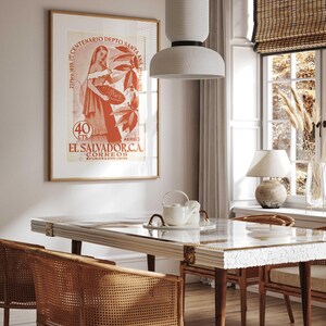 May include: A dining room scene featuring a rectangular table with a glass top and wooden legs, surrounded by woven chairs. A framed vintage poster with text in Spanish hangs on the wall. A white pendant light hangs above the table, and a window with a bamboo shade is visible.