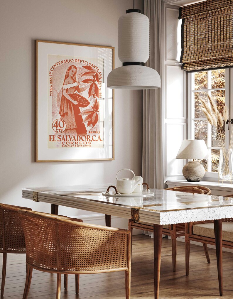 May include: A dining room scene featuring a rectangular table with a glass top and wooden legs, surrounded by woven chairs. A framed vintage poster with text in Spanish hangs on the wall. A white pendant light hangs above the table, and a window with a bamboo shade is visible.