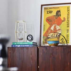 Pu&ograve; includere: Un armadio in legno con due ante, una clessidra dorata in una cornice geometrica, una pila di libri con i titoli "EPIC HIKES of the WORLD" e "WHERE TO GO WHEN", e un poster incorniciato con uno sfondo giallo e il testo "REPUBLIQUE du MALI" e "Kono: danse Bamanan 150f".