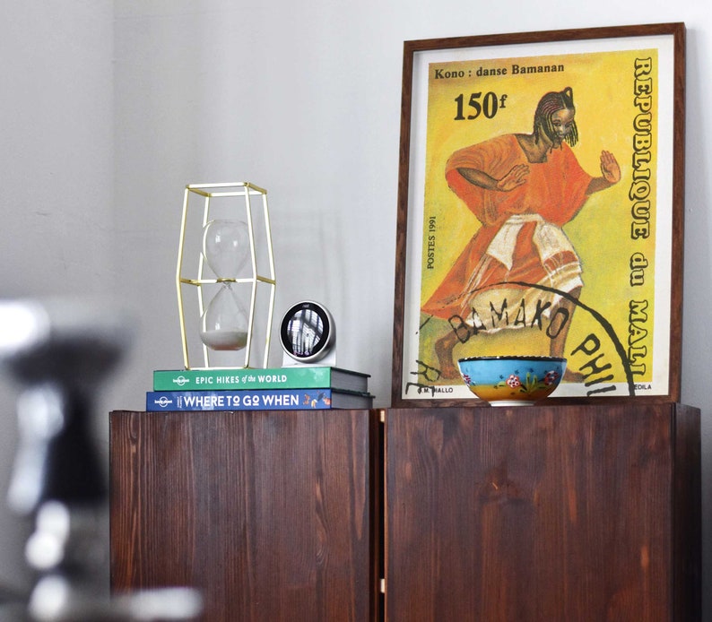 Pu&ograve; includere: Un armadio in legno con due ante, una clessidra dorata in una cornice geometrica, una pila di libri con i titoli "EPIC HIKES of the WORLD" e "WHERE TO GO WHEN", e un poster incorniciato con uno sfondo giallo e il testo "REPUBLIQUE du MALI" e "Kono: danse Bamanan 150f".