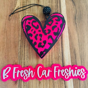 May include: Pink heart-shaped car freshener with a black leopard print design. The freshener is hanging from a black string with a black rhinestone embellishment. The text "B Fresh Car Freshies" is printed on a pink banner.