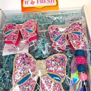 May include: Three pink glitter bow car air fresheners in a gift box. The bows have gold glitter and blue accents. The largest bow has a metal ring for hanging. The box is filled with blue shredded paper. The box has a label that says "B Fresh Car Fresheners".
