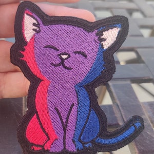 May include: A purple, red, and blue embroidered cat patch with a smiling face. The cat is sitting with its tail curled around its legs.