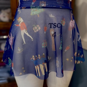 May include: A blue mesh ballet skirt with a festive print featuring nutcrackers, clocks, and wrapped gifts. The skirt has the text "TSC 4" printed on it. The skirt is wrapped around a mannequin.