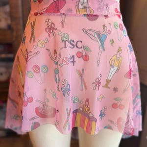 Puede incluir: Falda de ballet rosa con un estampado caprichoso de bailarinas, caramelos y cerezas. La falda tiene una capa de malla y presenta el texto "TSC 4".