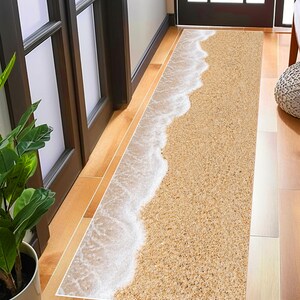 Peut inclure: Un tapis rectangulaire intérieur représentant une scène de plage. Le motif comprend une plage de sable avec des vagues blanches et mousseuses. Le tapis est placé dans une porte, avec une plante et un cadre de porte en bois en arrière-plan.