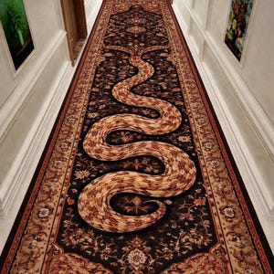 Peut inclure: Un long tapis rectangulaire avec un motif détaillé et un grand motif de serpent. Le tapis présente un fond sombre avec des motifs floraux et géométriques dans des tons de marron et de beige. Le serpent est de couleur marron clair avec un motif d'écailles.