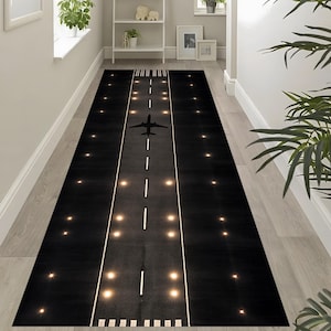May include: Black rectangular area rug with a runway design. The rug features a dark gray runway with white lines and a silhouette of an airplane. Yellow lights are scattered along the sides, creating a realistic airport scene.