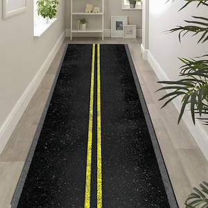 Könnte beinhalten: Ein langer, rechteckiger Teppich mit einem schwarzen Asphaltmuster und zwei parallelen gelben Linien in der Mitte. Der Teppich hat einen grauen Rand und liegt auf einem hellen Holzboden. Der Teppich ist als Straße gestaltet.
