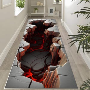 Op de afbeelding: Een rechthoekig gangtapijt met een 3D-afbeelding van een diepe kloof. Het ontwerp toont grijze rotsformaties met een vurige rode lava-achtige substantie aan de onderkant. Het tapijt is ontworpen om een optische illusie te creëren.