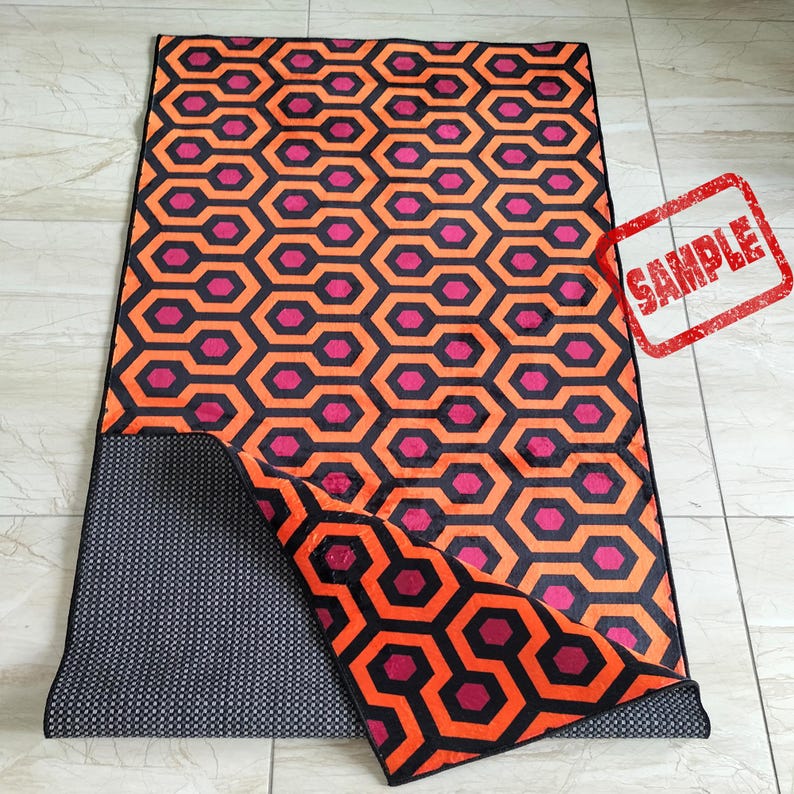 May include: A rectangular rug with a repeating geometric pattern of orange, black, and magenta hexagons. The rug is partially folded, revealing a dark gray, dotted underside. A red "SAMPLE" stamp is visible in the upper right corner.