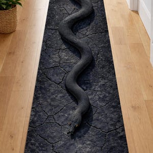 Puede incluir: Una alfombra larga y rectangular con una serpiente gris oscura detallada que serpentea sobre una superficie texturizada y agrietada. La cabeza de la serpiente está en la parte inferior, con ojos anaranjados. La alfombra está colocada sobre un suelo de madera.