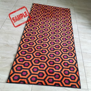 May include: A rectangular rug with a repeating geometric pattern of orange, black, and magenta hexagons. The rug has a black border and is displayed on a tiled floor. The word "SAMPLE" is stamped in red in the upper left corner.