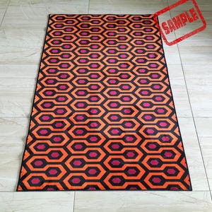May include: A rectangular rug with a repeating geometric pattern in orange, black, and magenta. The pattern consists of interconnected hexagons and elongated shapes. The word "SAMPLE" is stamped in red in the upper right corner.