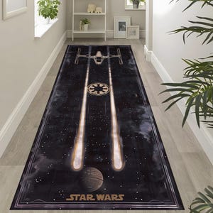 Puede incluir: Una alfombra rectangular con temática de Star Wars con un fondo negro que presenta una nave espacial, un símbolo y dos rayos de luz. La alfombra también muestra una Estrella de la Muerte y las palabras "STAR WARS" en dorado.