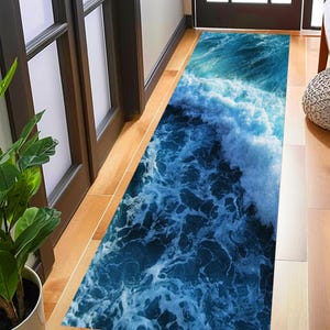 Puede incluir: Una alfombra rectangular con un diseño dinámico de olas oceánicas. La alfombra presenta tonos de azul y blanco, que representan un patrón de olas. La alfombra está colocada sobre un suelo de madera, añadiendo un toque de decoración costera al espacio.