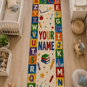 Può includere: Un tappeto rettangolare con un centro color crema che riporta il testo YOUR NAME, circondato da un bordo di blocchi alfabetici colorati. Il design include illustrazioni di libri, mele, matite, pastelli e stelle sulla superficie del tappeto.