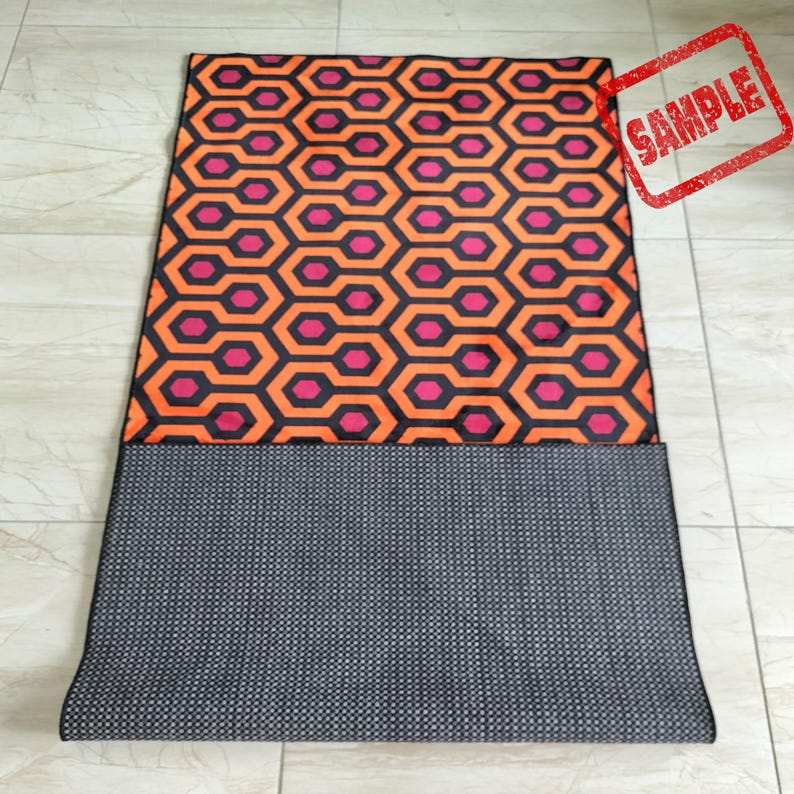 May include: A rectangular rug with a repeating geometric pattern of orange, pink, and black hexagons. The bottom portion of the rug is a dark gray, textured material. A red stamp with the word "SAMPLE" is in the upper right corner.
