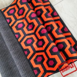 May include: A folded rug with a geometric pattern in black, orange, and magenta. The design features interlocking hexagons. A section of the rug has a gray and white dotted pattern. A red "SAMPLE" stamp is visible.