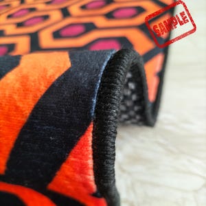 May include: Close-up of a rug with a repeating geometric pattern in orange, black, and pink. The rug's edge is visible, showing a black border. The word "SAMPLE" is stamped on the rug.