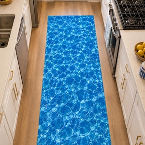 May include: A rectangular kitchen runner rug with a vibrant blue water ripple design. The rug is placed on a light wood floor between white cabinets and a stainless steel appliance. The rug's dimensions are approximately 2.5 feet wide and 6 feet long.